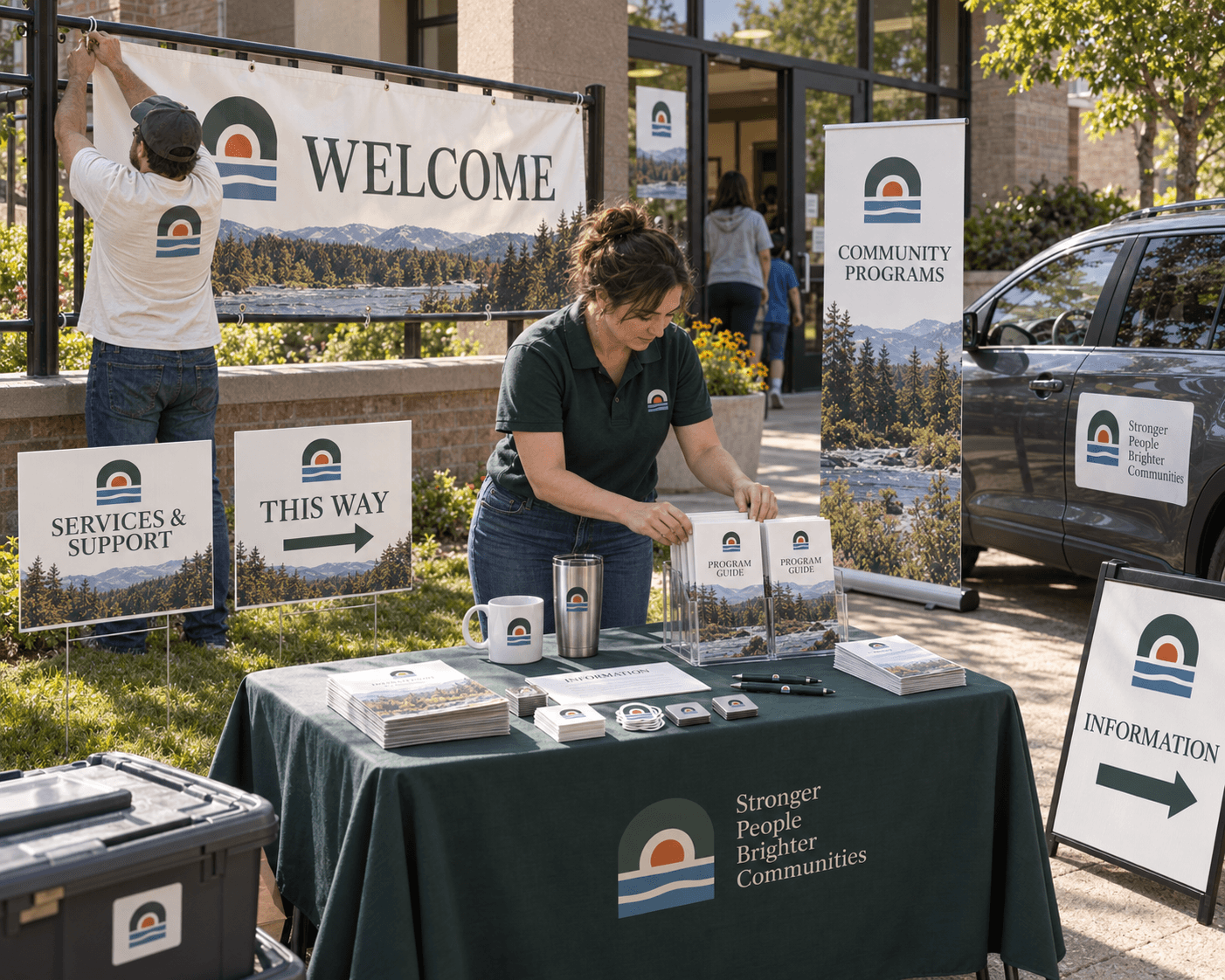 Volunteers setting up an outdoor welcome table with directional signs, a banner, and printed handouts.