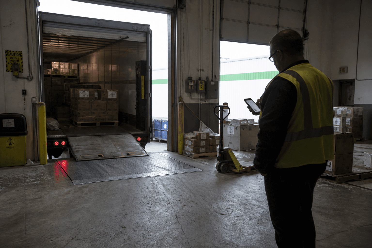 A worker in a high-visibility vest stands in a dim loading dock doorway checking a phone.